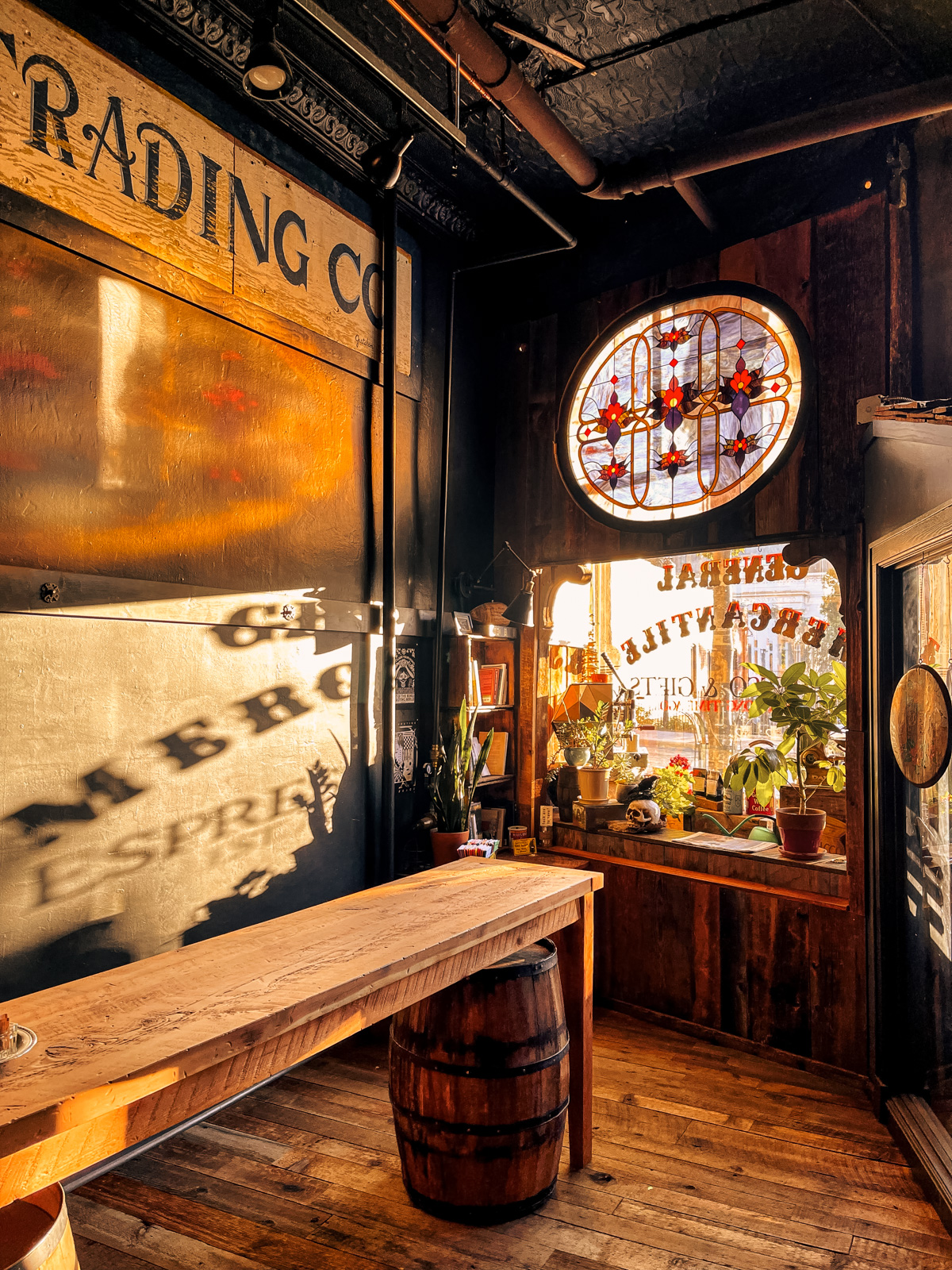 Late-afternoon sun across a reclaimed-wood counter and barrel inside the General Mercantile, with an oval stained-glass window above the front display